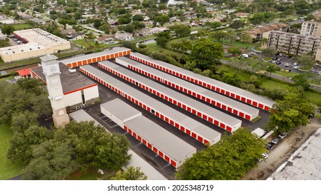 An Aerial View Over A Public Storage Facility With Long Rows Of Storage Units With Orange Garage Doors. There Is A Tall Clock Tower In Front. It Is Surrounded By Green Trees On A Sunny Day In Florida.