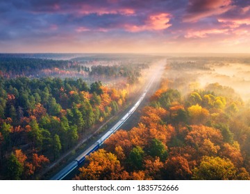 Aerial View Of Passenger Train And Beautiful Forest In Fog At Sunset. Autumn Landscape With Railroad, Moving Train, Foggy Trees And Colorful Sky With Clouds In Fall. Top View Of Railway Station