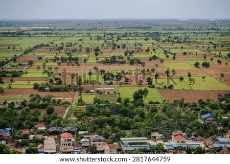 Aerial view of patchwork agricultural fields with scattered trees stretching behind a small village near Banteay Srei in rural Cambodia. Banteay Srei, Cambodia - June 11, 2023