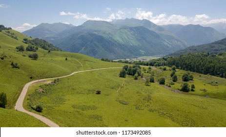 An Aerial View Of A Path Along Which Cyclists Cycle Through Green Fields And Mountains On A Sunny Day.