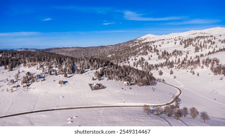 An aerial view of a peaceful snow-covered valley with scattered houses, surrounded by dense evergreen forests and rolling hills. The winding road cuts through the pristine winter landscape - Powered by Shutterstock