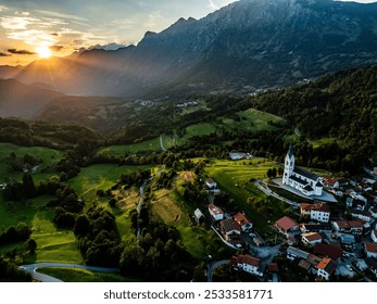 Aerial view of a picturesque village with a church surrounded by green fields and mountains at sunset. - Powered by Shutterstock