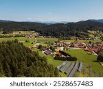 Aerial view of a picturesque village surrounded by lush green forests and hills. Solar panels are visible in the foreground, showcasing renewable energy use. The landscape features fields and scattere