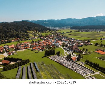 Aerial view of a picturesque village surrounded by lush green fields and mountains. The village features traditional houses with red roofs, a cemetery, and solar panels in the foreground. - Powered by Shutterstock