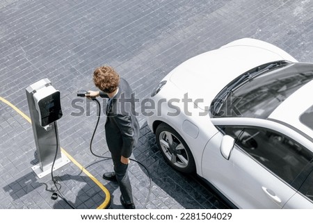 Aerial view of progressive businessman in black formal suit with his electric vehicle recharging battery at public car park charging station as vehicle powered by sustainable energy concept.