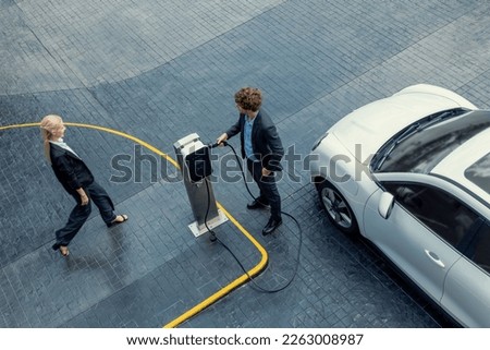 Aerial view progressive lifestyle of businesspeople with electric car connected to charging station at city center public parking car. Eco friendly rechargeable car powered by alternative clean energy