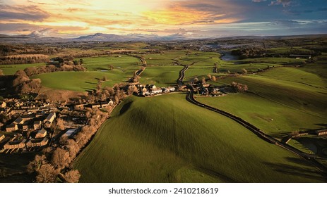 Aerial view of a quaint village amidst lush green fields with rolling hills in the background during sunset. - Powered by Shutterstock