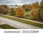 Aerial view of a quiet city street next to a small park with bright autumn trees and green lawns. A white building stands in the background under a cloudy sky, paths, garden beds and benches can be se