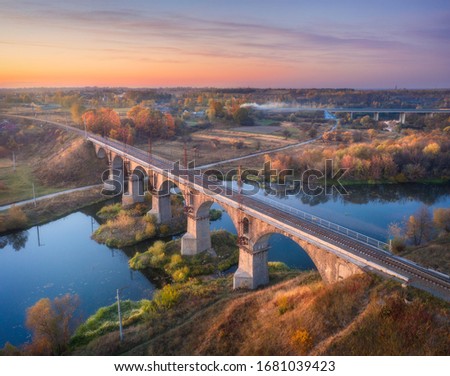 Aerial view of railroad bridge and river at beautiful sunset in autumn. Industrial landscape with bridge, blue water, colorful trees, rural railway station, orange sky in fall at dusk. Top view