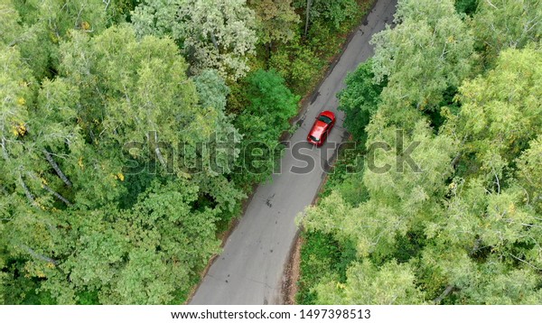 Aerial view of red car\
driving on country road in forest. Shot from drone flying over road\
in autumn forest