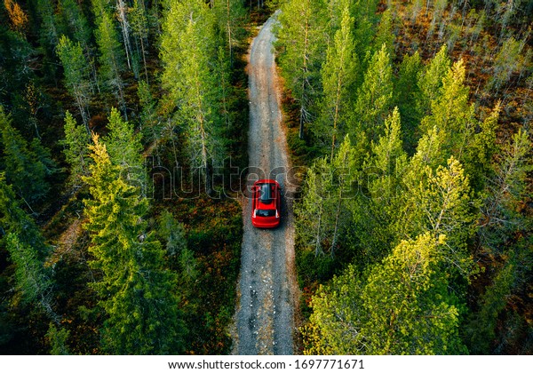 aerial view of a red car driving on a gravel road through the forest.