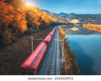 Aerial view of red freight train moving near river in alpine mountains at sunrise in autumn. Top view of wagons, railroad, lake, reflection in water, orange trees in fall. Railway station in Slovenia - Powered by Shutterstock