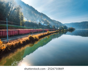 Aerial view of red freight train moving near lake in alpine mountains at sunrise. Autumn. Top view of wagons, sunbeams, railroad, river, reflection in water, orange trees in fall. Railway in Slovenia - Powered by Shutterstock