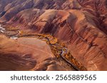 Aerial view of red and orange hills with a curving river lined by yellow autumn trees. Vibrant natural scenery showcasing rugged erosion patterns in a dramatic and vibrant environment.