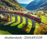 Aerial view of red train on Kreisviadukt in alpine mountains at sunset in autumn. Bernina Express, Switzerland. Top view of train, railroad, green grass , colorful trees in fall. Brusio spiral viaduct