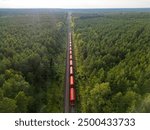 Aerial view of a red train transporting materials on tracks through a coniferous forest in Europe, delivery and transportation of goods, economic development, import, export, and railway transport.