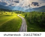 Aerial view of road in green meadows at sunset in summer. Top view from drone of rural road, mountains, forest. Beautiful landscape with roadway, sun rays, trees, hills, green grass, clouds. Slovenia