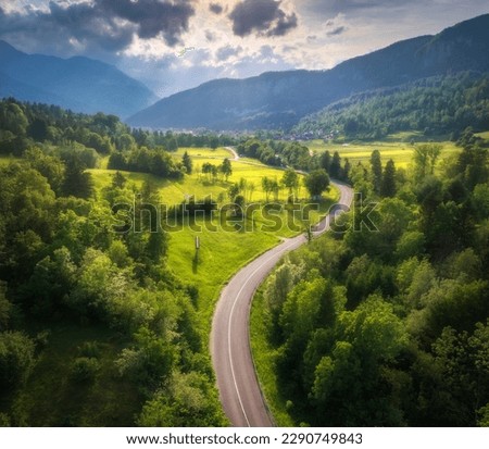 Aerial view of road in green meadows at summer sunny day. Top view from drone of rural road, mountains, forest. Beautiful landscape with roadway, sun rays, trees, hills, green grass, clouds. Slovenia