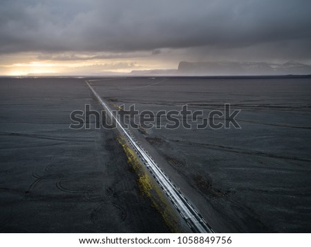 Aerial view of a road leading across vast open land area with black volcanic sand and mountains in the distance and golden sunset behind cloudy sky in Haoldukvisl, Iceland