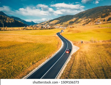 Aerial view of the road in mountain valley at sunset in autumn. Top view of asphalt roadway, car, hills with orange grass, blue sky, trees, buildings. Highway and fields in fall. Colorful landscape