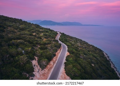 Aerial View Of Road Near Blue Sea, Forest At Sunset In Summer. Travel In Lefkada, Greece. Top View Of Beautiful Road, Pink Sky, Green Trees. Colorful Landscape With Highway And Sea Coast. Road Trip