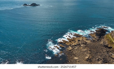 Aerial view of a rocky coastline with clear blue waters and gentle waves. An isolated rocky island is visible in the distance, surrounded by the ocean. At Land's End, Cornwall, UK. - Powered by Shutterstock