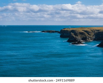 Aerial view of a rocky coastline in Iceland with calm blue ocean, green cliffs, and small white houses in the distance under a bright blue sky. - Powered by Shutterstock