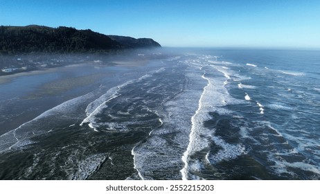 Aerial view of rolling ocean waves crashing onto a wide sandy shore with misty hills in the background, creating a dramatic coastal scene under a clear blue sky. - Powered by Shutterstock