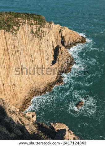Aerial view of a rugged cliff with vertical striations, jutting into a turbulent sea with white waves crashing against it.