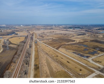 Aerial view of rural farm land with railroad tracks running through, dirt roads, blue sky, puddling water from melting snow. Trees along the railroad tracks. Transmission towers in the farm fields.  