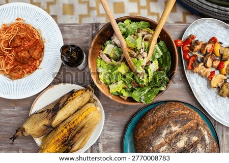 Aerial view of a rustic table with grilled corn, fresh salad, spaghetti with meatballs, skewers, and crusty bread, alongside a bottle of beer.