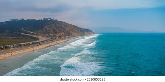 Aerial view of San Diego's coastal area featuring a sandy beach, Pacific Ocean, rugged hills, and a road with parked vehicles and beachgoers. - Powered by Shutterstock