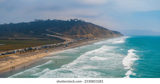 Aerial view of San Diego's coastline featuring parked vehicles along a road, gentle waves on the sandy shore, and a rugged hill under a partly cloudy sky. - Powered by Shutterstock