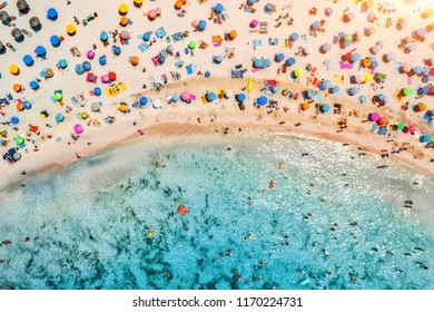 Aerial view of sandy beach with colorful umbrellas, swimming people in sea bay with transparent blue water at sunset in summer. Travel in Mallorca, Balearic islands, Spain. Top view. Landscape