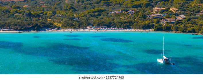 Aerial view of a Sardinian beach with turquoise waters, a sailboat, sunbathers, umbrellas, and villas nestled in lush greenery. - Powered by Shutterstock