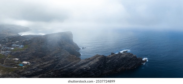 Aerial view of Sardinia's coastline featuring steep cliffs, mist, and a small settlement with buildings and a swimming pool on the cliffside. - Powered by Shutterstock