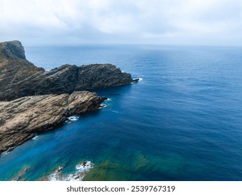 Aerial view of Sardinia's coastline featuring rocky cliffs and a gradient of turquoise to deep blue sea under an overcast sky. - Powered by Shutterstock