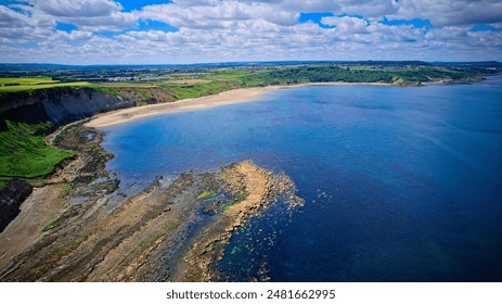 Aerial view of a scenic coastline with clear blue water, rocky shore, and green fields under a partly cloudy sky. - Powered by Shutterstock