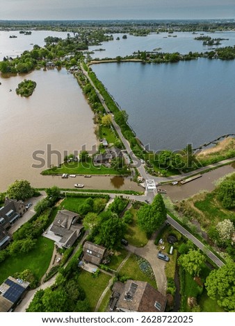 Aerial view of a scenic lakeside village with narrow peninsulas, green fields, and houses surrounded by calm waters under a cloudy sky in the Dutch countryside.