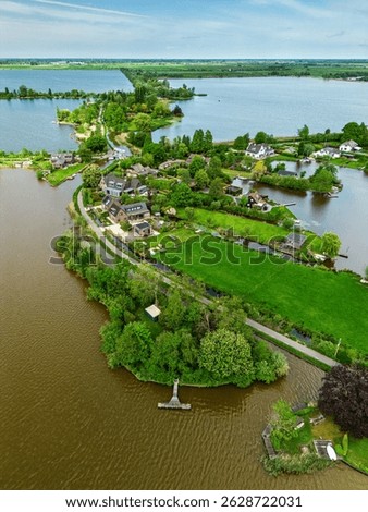 Aerial view of a scenic lakeside village with narrow peninsulas, green fields, and houses surrounded by calm waters under a cloudy sky in the Dutch countryside.