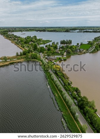 Aerial view of a scenic lakeside village with narrow peninsulas, green fields, and houses surrounded by calm waters under a cloudy sky in the Dutch countryside.