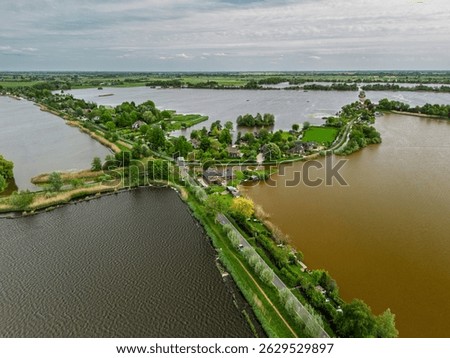 Aerial view of a scenic lakeside village with narrow peninsulas, green fields, and houses surrounded by calm waters under a cloudy sky in the Dutch countryside.