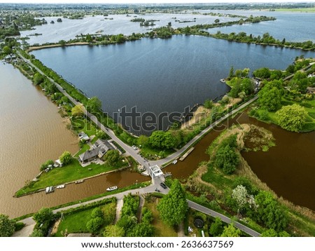 Aerial view of a scenic lakeside village with narrow peninsulas, green fields, and houses surrounded by calm waters under a cloudy sky in the Dutch countryside.