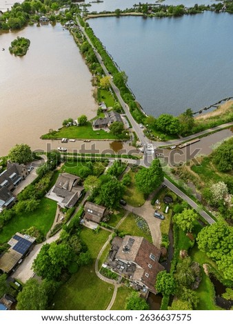 Aerial view of a scenic lakeside village with narrow peninsulas, green fields, and houses surrounded by calm waters under a cloudy sky in the Dutch countryside.
