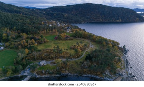 Aerial view of a scenic landscape featuring a coastline, lush green fields, and a forested hillside. The image captures a tranquil bay with a few houses nestled among trees, showcasing autumn colors. - Powered by Shutterstock