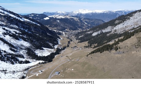 Aerial view of a scenic mountain valley in early spring, featuring snow-capped peaks, grassy slopes, winding roads, and scattered cabins - Powered by Shutterstock