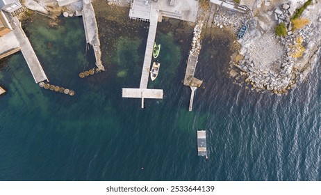 An aerial view of a serene coastal area featuring wooden docks extending into clear blue waters. Several boats are moored alongside the docks, with rocky shorelines and scattered boulders visible. - Powered by Shutterstock