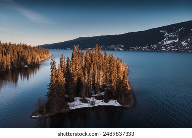 An aerial view of a serene lake with a small, tree-covered island in the center. The tranquil water reflects the surrounding forested hills and distant mountains, partially covered in snow. The late a - Powered by Shutterstock