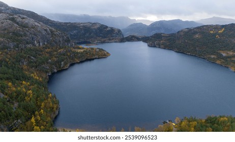 Aerial view of a serene lake surrounded by rugged mountains and autumn foliage. The landscape features a mix of green and yellow trees, with rocky cliffs and a cloudy sky in the background. - Powered by Shutterstock