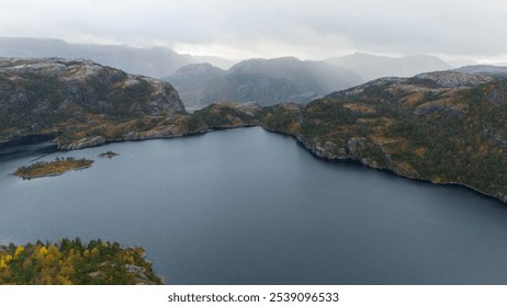 Aerial view of a serene lake surrounded by rugged mountains and autumn foliage. The landscape features a mix of green and yellow trees, with rocky cliffs and a cloudy sky in the background. - Powered by Shutterstock